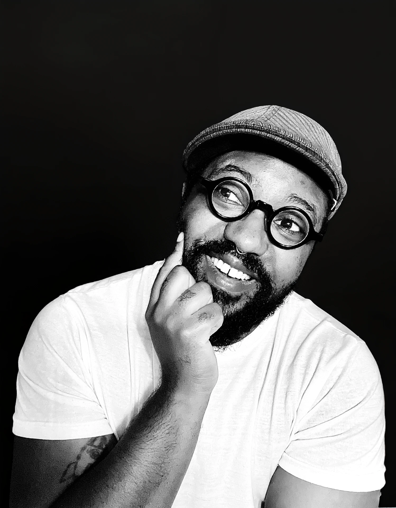 A black and white photo of Taj M. Smith, a bearded Black man wearing a flat cap, round black glasses, and a white t-shirt. He smiles, looking playfully up to the right with a hand on his chin. 